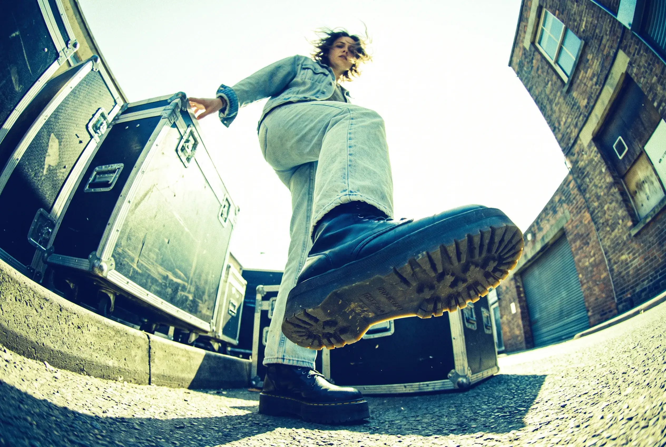 Hero photograph: a worm's-eye view of a model standing over the camera on stacked road cases, one platform boot filling the lower third of the frame against a blown midday sky.
