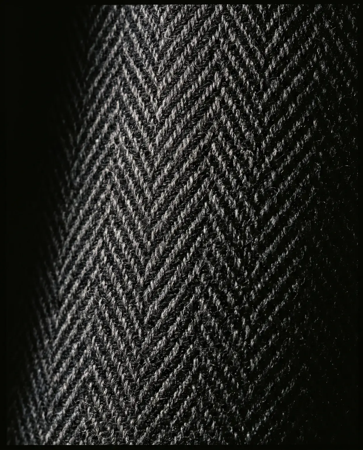 Macro of charcoal herringbone wool, raking light throwing a shadow off every chevron