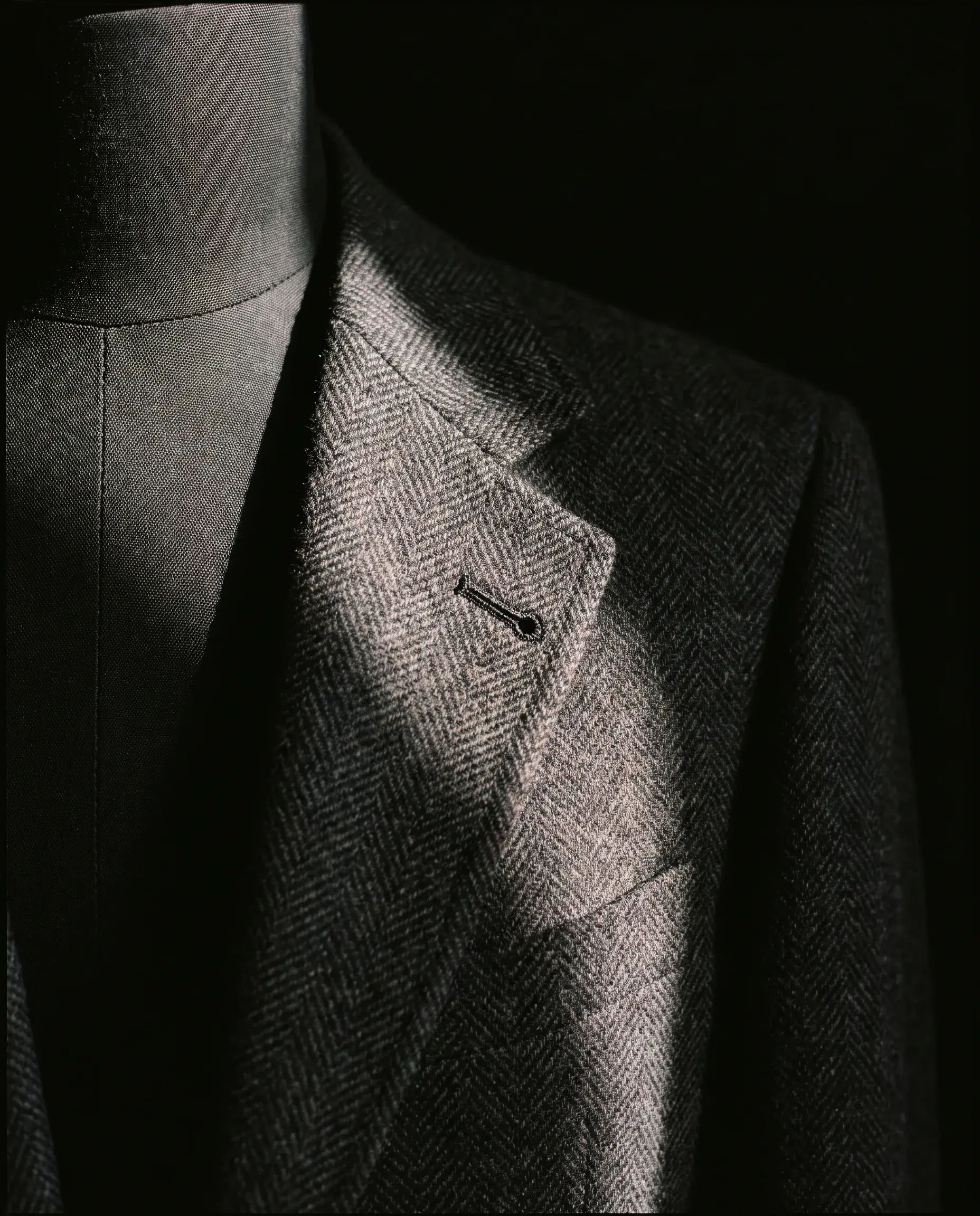The lapel of the finished jacket in near darkness, a raking light running along the belly of its roll and catching the hand-sewn buttonhole