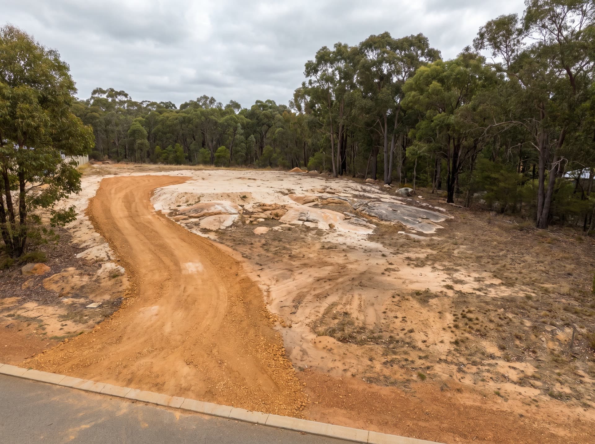 Cleared sloping block seen from the street: a raw orange laterite driveway cut curves up from the verge, exposed granite sheet rock beside it, jarrah and marri along the back boundary under an overcast sky